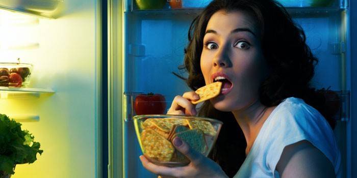 Девушка перед открытым холодильником ест крекеры