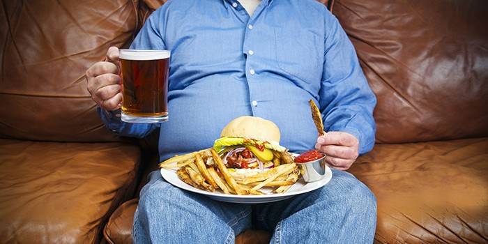 Полный мужчина с кружкой пива и вредной пищей сидит на диване
